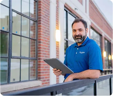 A person stands outside a building holding a tablet. The setting features brick walls and large windows, with soft outdoor lighting.