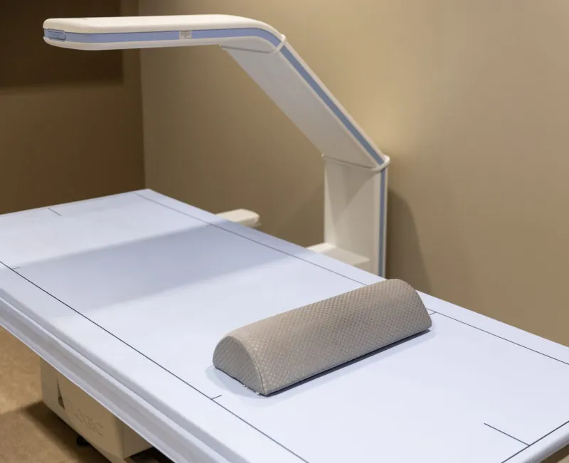 A medical examination table with a curved support cushion is positioned under a ceiling-mounted light. The setting appears to be a clinical or healthcare environment.