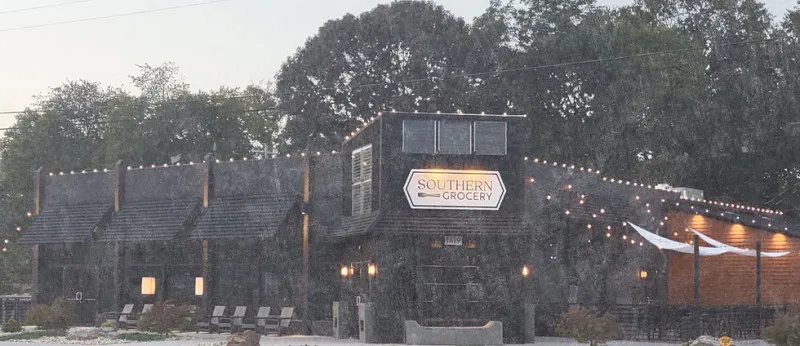 A grocery store building with a sign reading "SOUTHERN GROCERY" is illuminated by string lights. The setting features trees and outdoor seating in front.