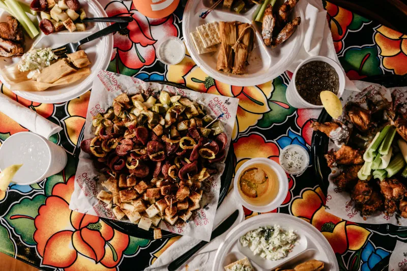 A table is covered with colorful floral tablecloths and filled with various dishes, including platters of meats, vegetables, and sauces, alongside drinks in cups.