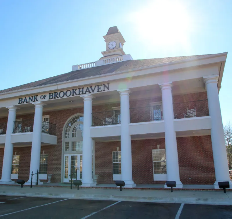 The exterior of a brick bank building features white columns and a clock tower. The words "BANK OF BROOKHAVEN" are prominently displayed above the entrance. Sunlight is visible in the background.