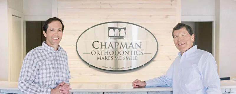 Two people are standing behind a reception desk in an office. A sign on the wall reads "Chapman Orthodontics Makes Me Smile," along with a website URL.
